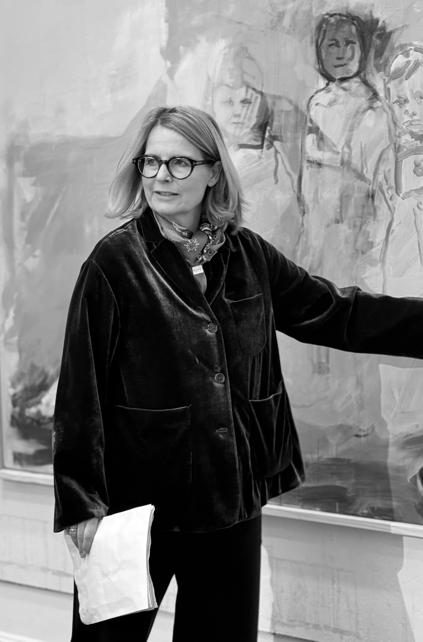 Schwarzweißfoto von Susan Hagemann mit Brille und Samtjacke, die vor einem großformatigen Gemälde den Arm ausstreckt