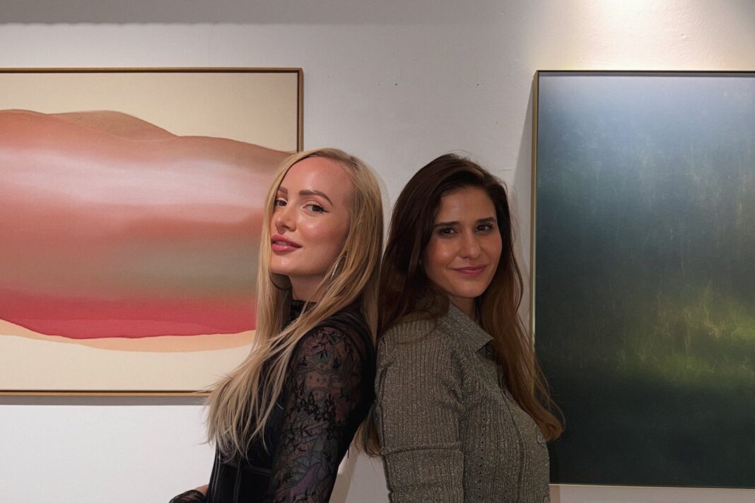 Shelly Thanner und Ulrike Pichl in der Galerie Ostermayr Zwei Künstlerinnen stehen Rücken an Rücken vor ihren Werken: links Shelly Thanner mit langen blonden Haaren, rechts Ulrike Pichl mit langen dunklen Haaren, dahinter ein rosa-rotes abstraktes Gemälde und eine dunkelgrüne Fotografie