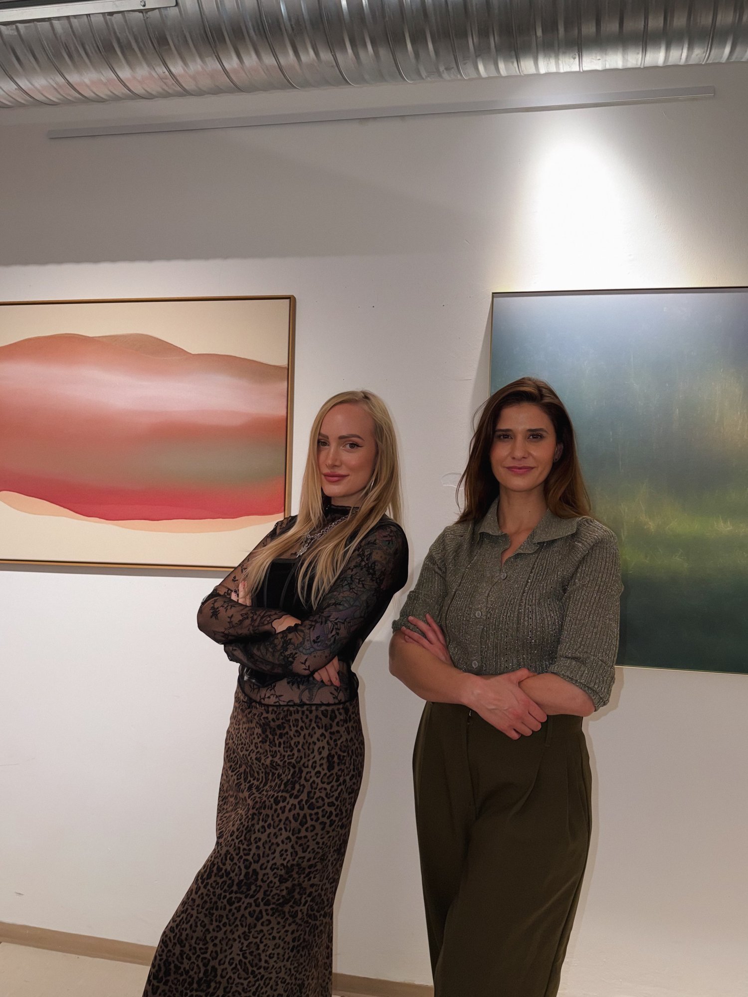 Shelly Thanner und Ulrike Pichl stehen mit verschränkten Armen nebeneinander in der Galerie, links hängt ein rosa-rotes abstraktes Gemälde, rechts eine dunkelgrüne Fotografie