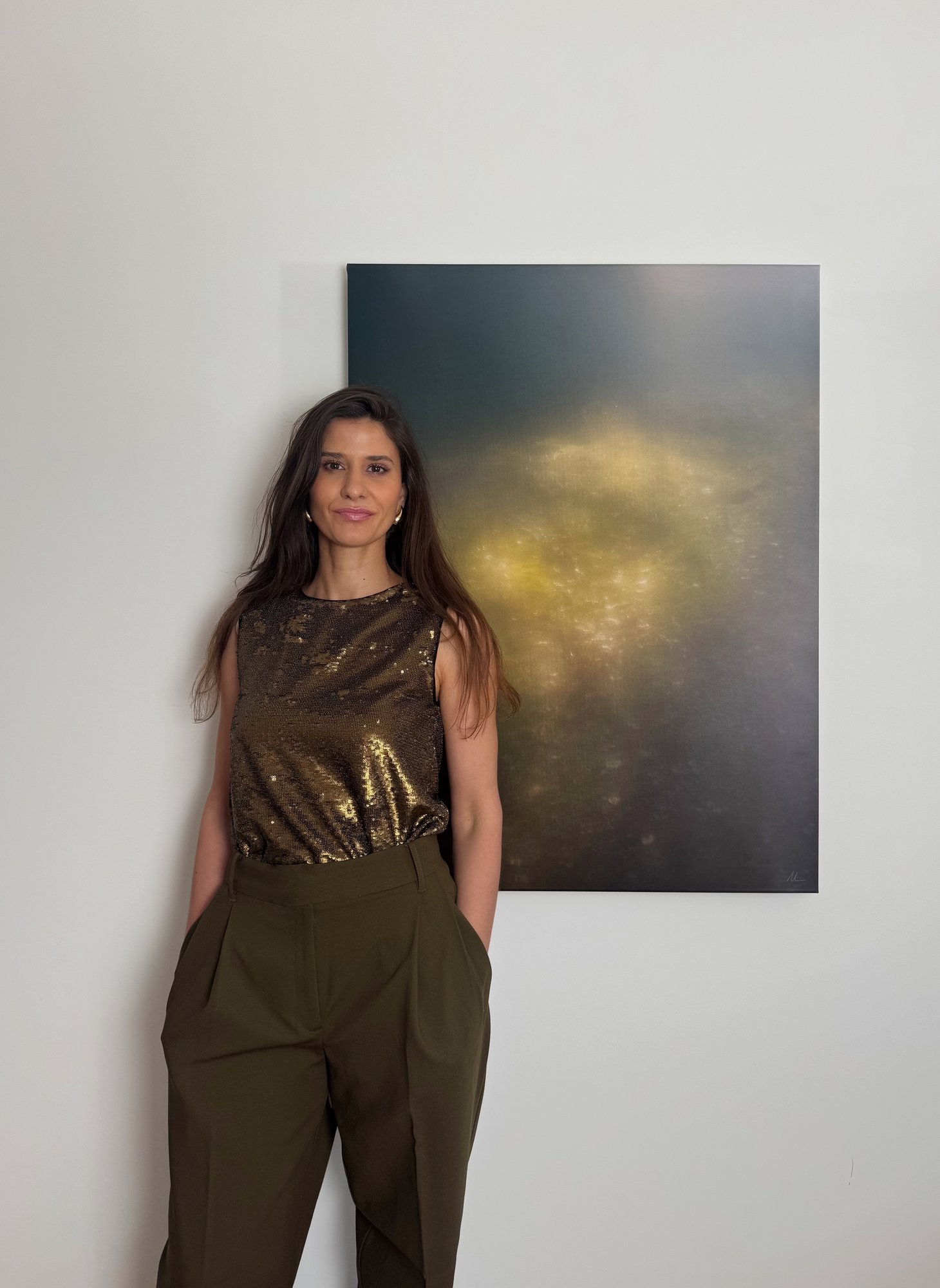 Ulrike Pichl in goldglänzendem Top und olivgrüner Hose steht an einer weißen Wand neben ihrer Fotografie mit gelb leuchtenden Lichtpunkten auf dunklem Grund