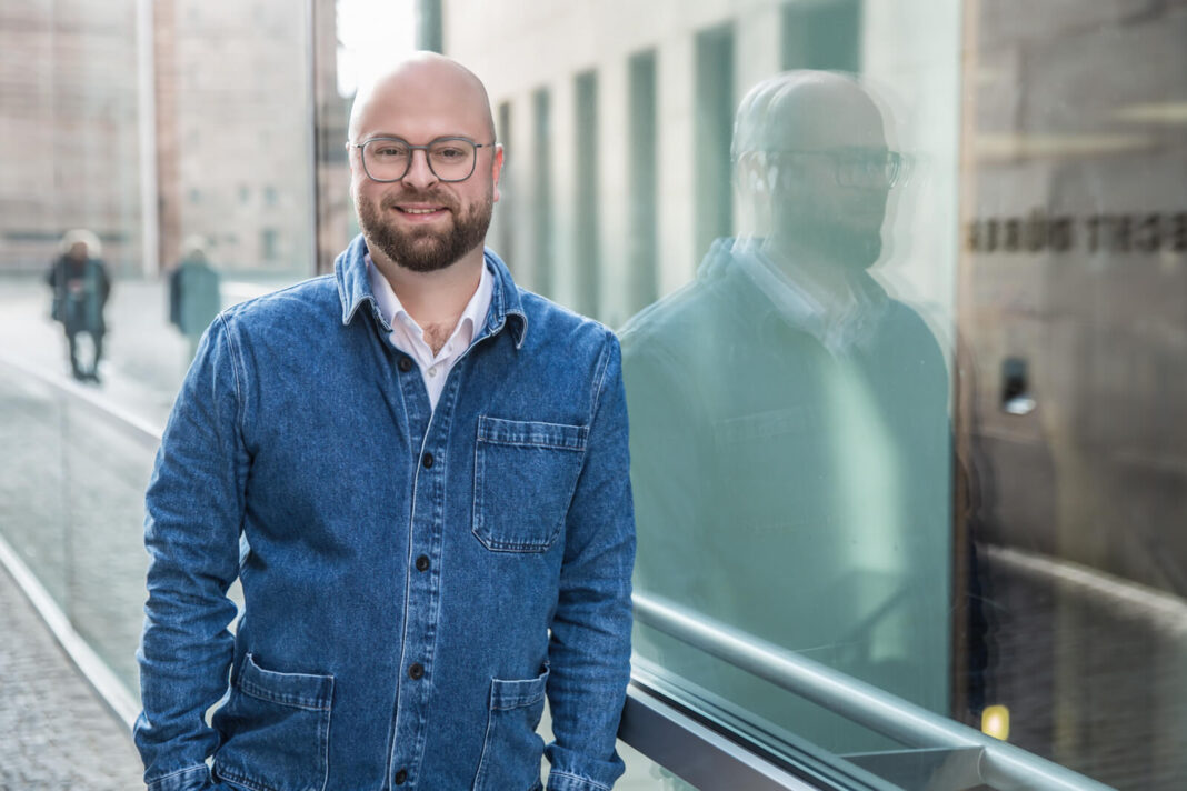 Dr. Alexander Rácz, ein Mann mit Glatze, Brille und Bart in Jeansjacke und weißem Hemd, lächelt in die Kamera und lehnt an einer Glasfläche, in der er sich spiegelt, dahinter eine Straße in Nürnberg